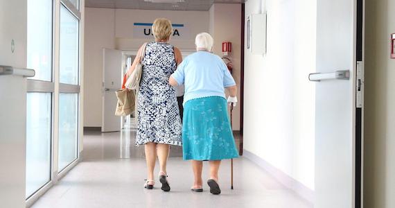 Older woman walking with cane down hospital hallway with younger woman